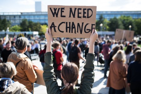 Scene from a protest, hundreds of people. A person with long brown hair in a ponytail holds a cardboard sign with black handwriting: "We need a change".